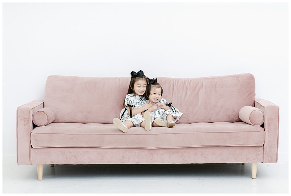 Sisters hugging on light pink sofa at The Lumen Room in Plano, TX photographed by Dallas family photographer Jenny Bui of Picture Bouquet Studio. 
