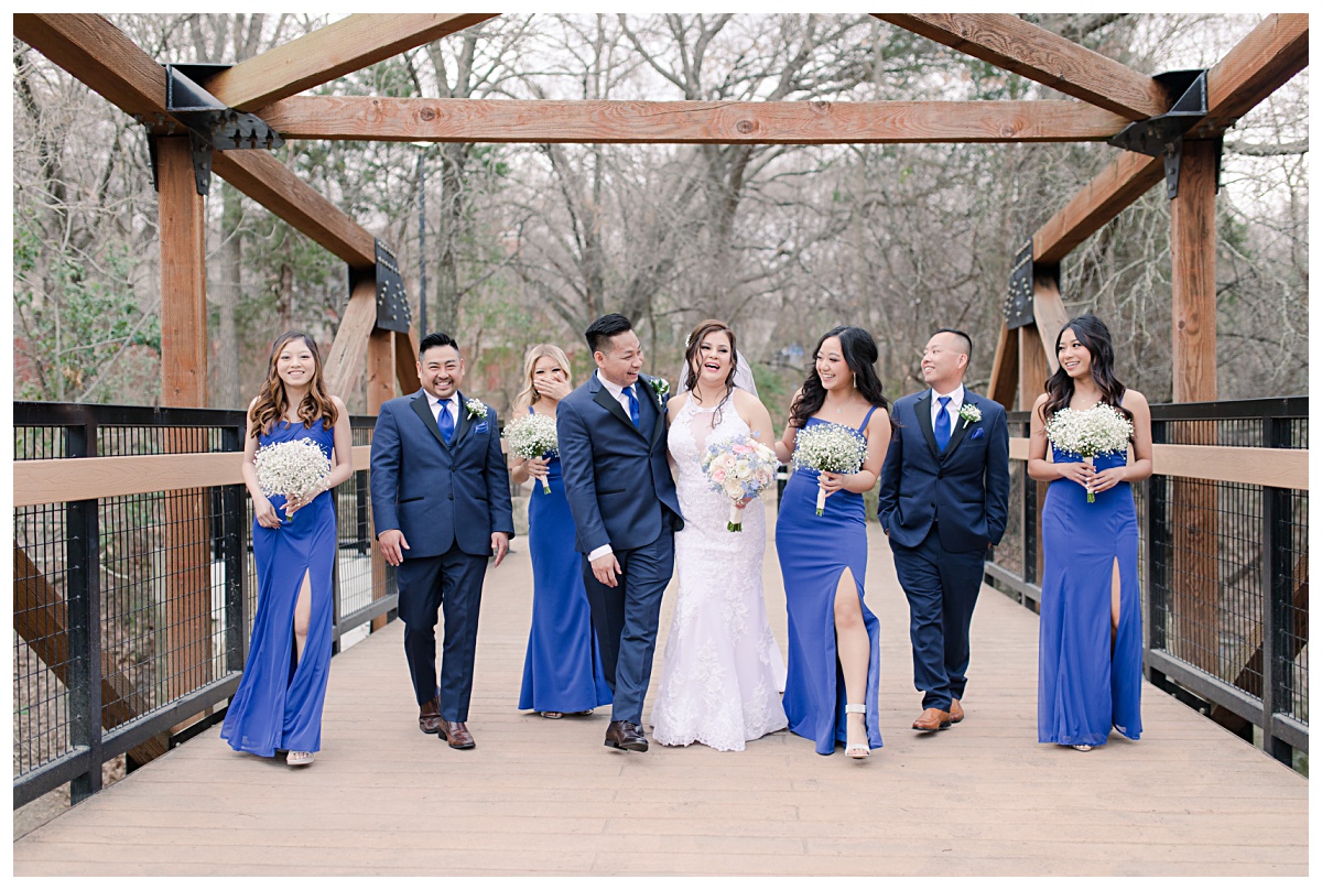 Bridal party in royal blue ensemble laughing as they walk on bridge at Praire Creek Park in Richardson, TX for bridal party photos for Dallas Vietnamese wedding photographed by Jenny Bui of Picture Bouquet Studio. 