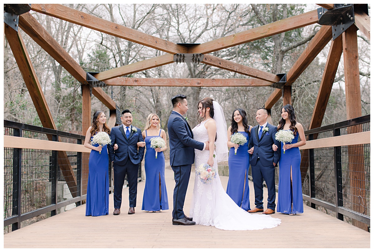 Bridal party in royal blue ensemble looking at bride and groom on bridge at Praire Creek Park in Richardson, TX for bridal party photos for Dallas Vietnamese wedding photographed by Jenny Bui of Picture Bouquet Studio. 