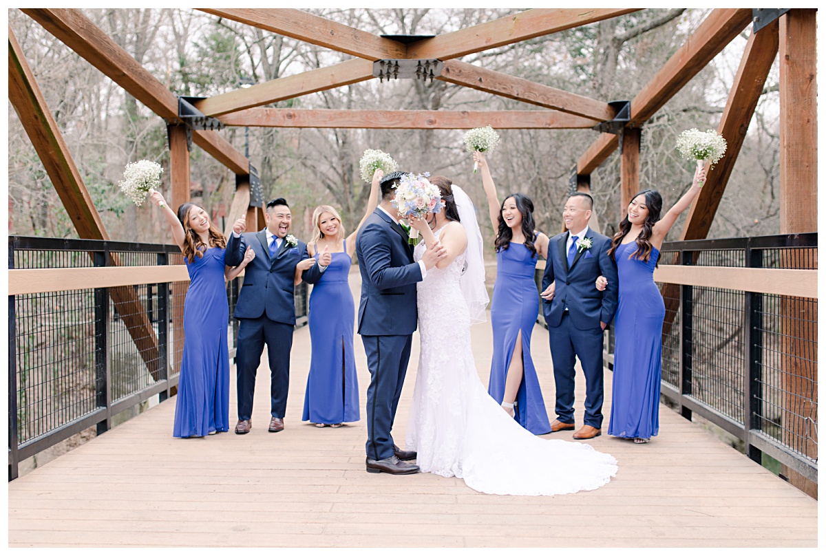 Royal blue bridal party cheers as bride and groom kiss behind bouquet on bridge at Prairie Creek Park in Richardson, TX for Dallas Vietnamese wedding photographed by Jenny Bui of Picture Bouquet Studio. 