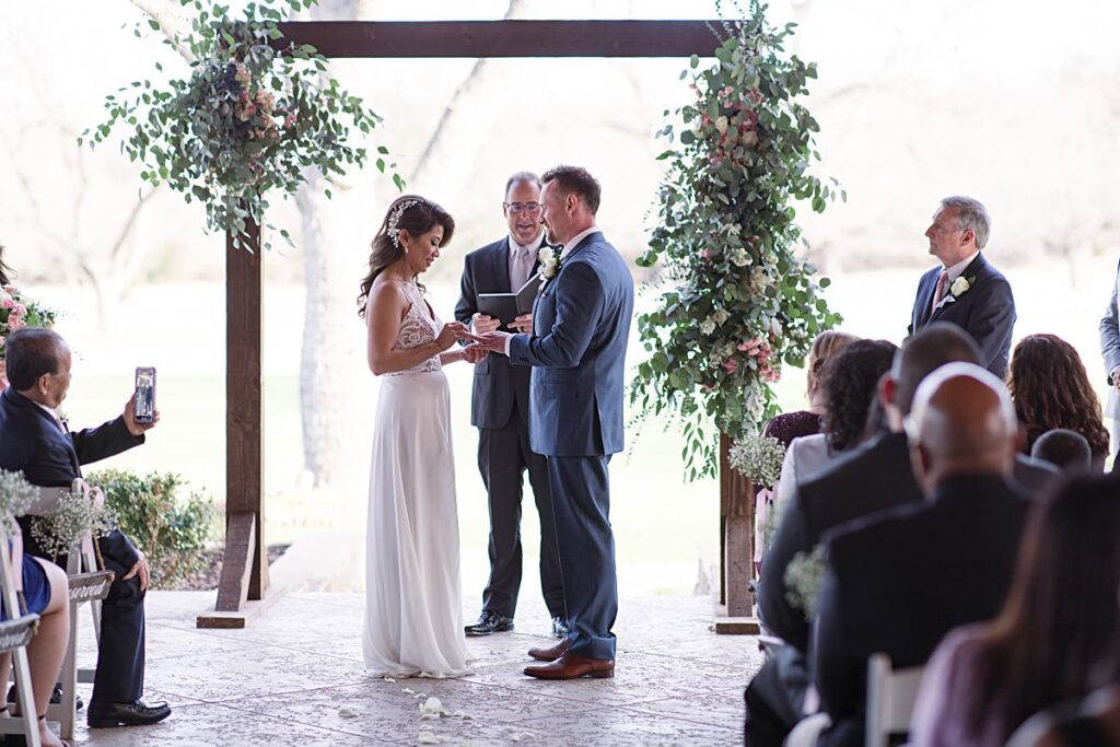 Bride and groom in front of floral arch at The Orchard Wedding Venue exchanging rings during their outdoor wedding in Texas photographed by Dallas Wedding Photographer Picture Bouquet Studio. 
