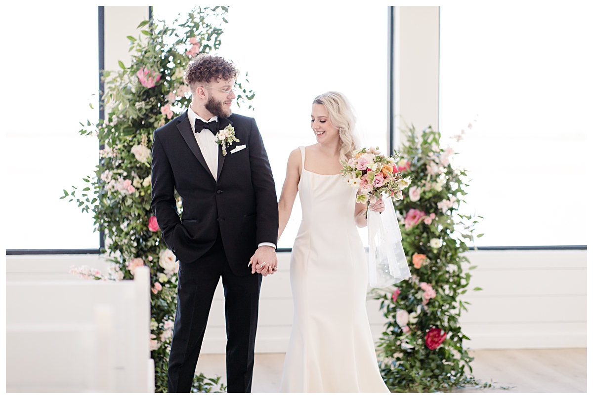 Bride and groom smiling at one another in front of floral arches in all white chapel at The Gardenia Venue photographed by Jenny Bui of Picture Bouquet Studio.