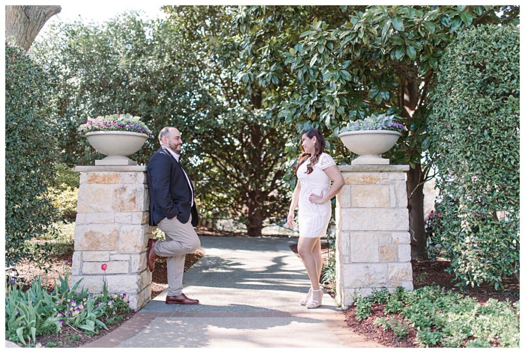 Couple in dressy attire stands against column gazing at one another from afar  for a Dallas spring engagement session at the Dallas Arboretum photographed by Jenny Bui of Picture Bouquet Studio. 