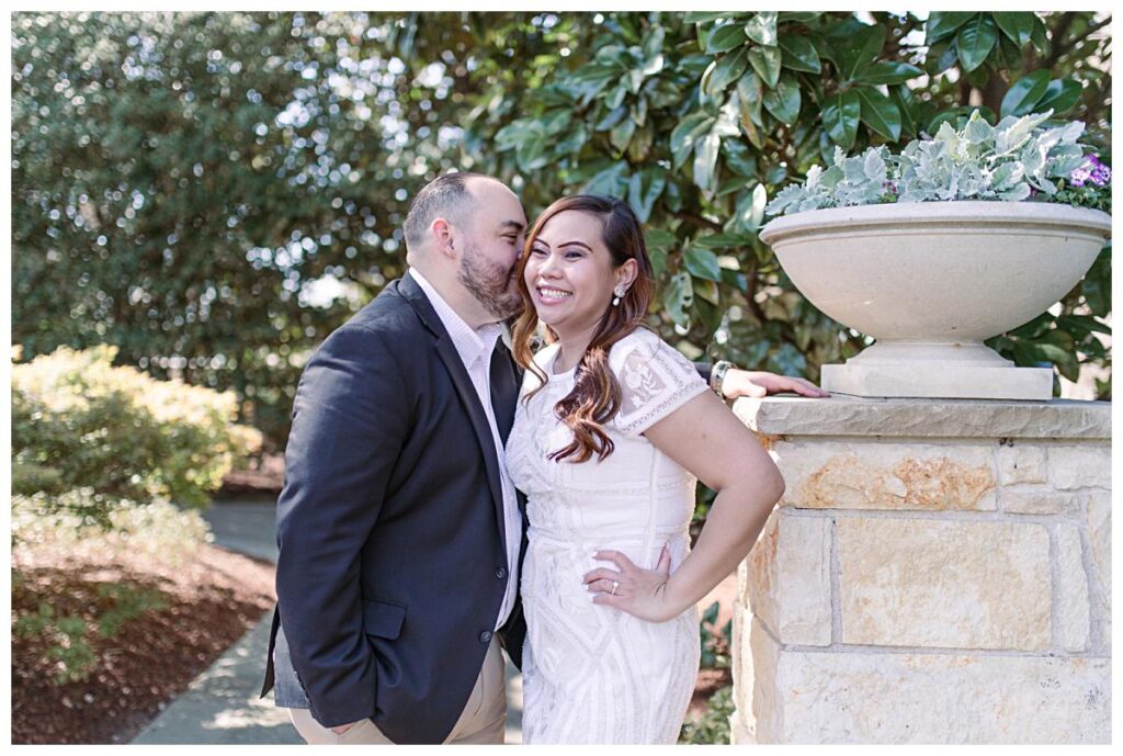 Guy in dark blazer leans and whispers into his fiancee's ears during a Dallas spring engagement session at the Dallas Arboretum photographed by Jenny Bui of Picture Bouquet Studio. 