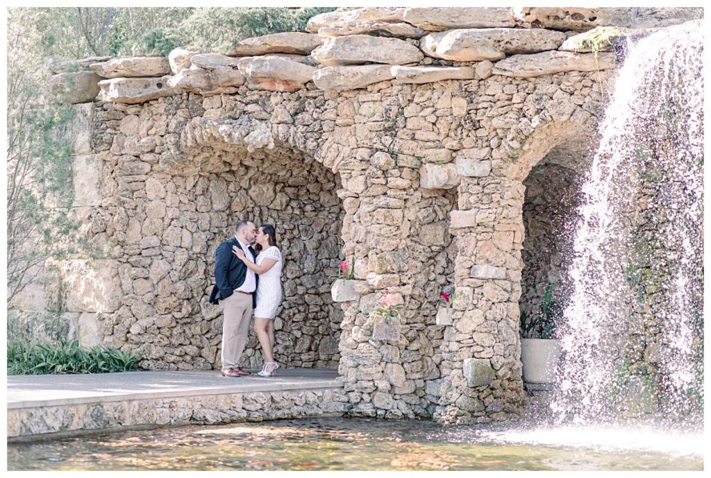 Engaged couple shares a kiss by the waterfall at the Dallas Arboretum for their spring engagement session photographed by Jenny Bui of Picture Bouquet Studio. 