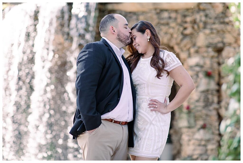 Guy in black blazer kisses fiancee's forehead by the waterfall at the Dallas Arboretum for their spring engagement session photographed by Jenny Bui of Picture Bouquet Studio. 