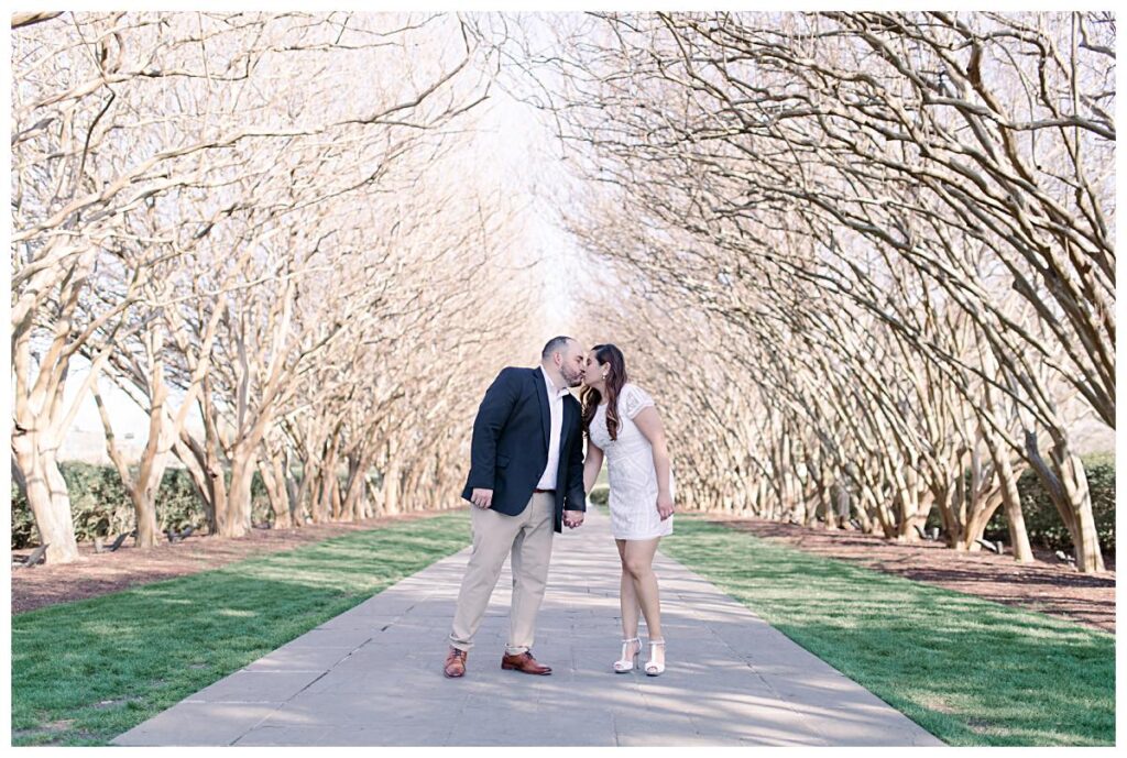 Engaged couple shares a kiss underneath the tunnel of crepe myrtle trees at the Dallas Arboretum for their spring engagement session photographed by Jenny Bui of Picture Bouquet Studio. 
