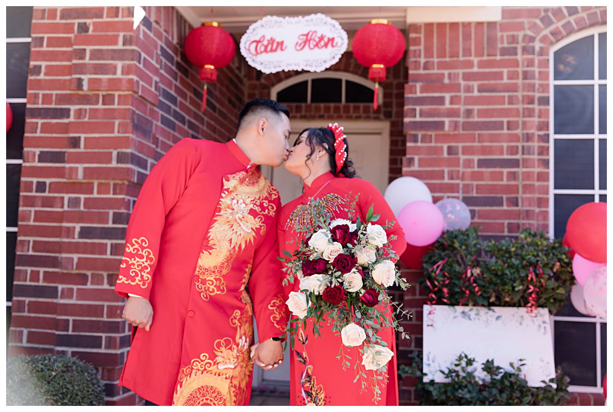 Vietnamese groom and bride in traditional red ao dais kiss under Tan Hon sign for Dallas Vietnamese tea ceremony photographed by Jenny Bui of Picture Bouquet Studio. 
