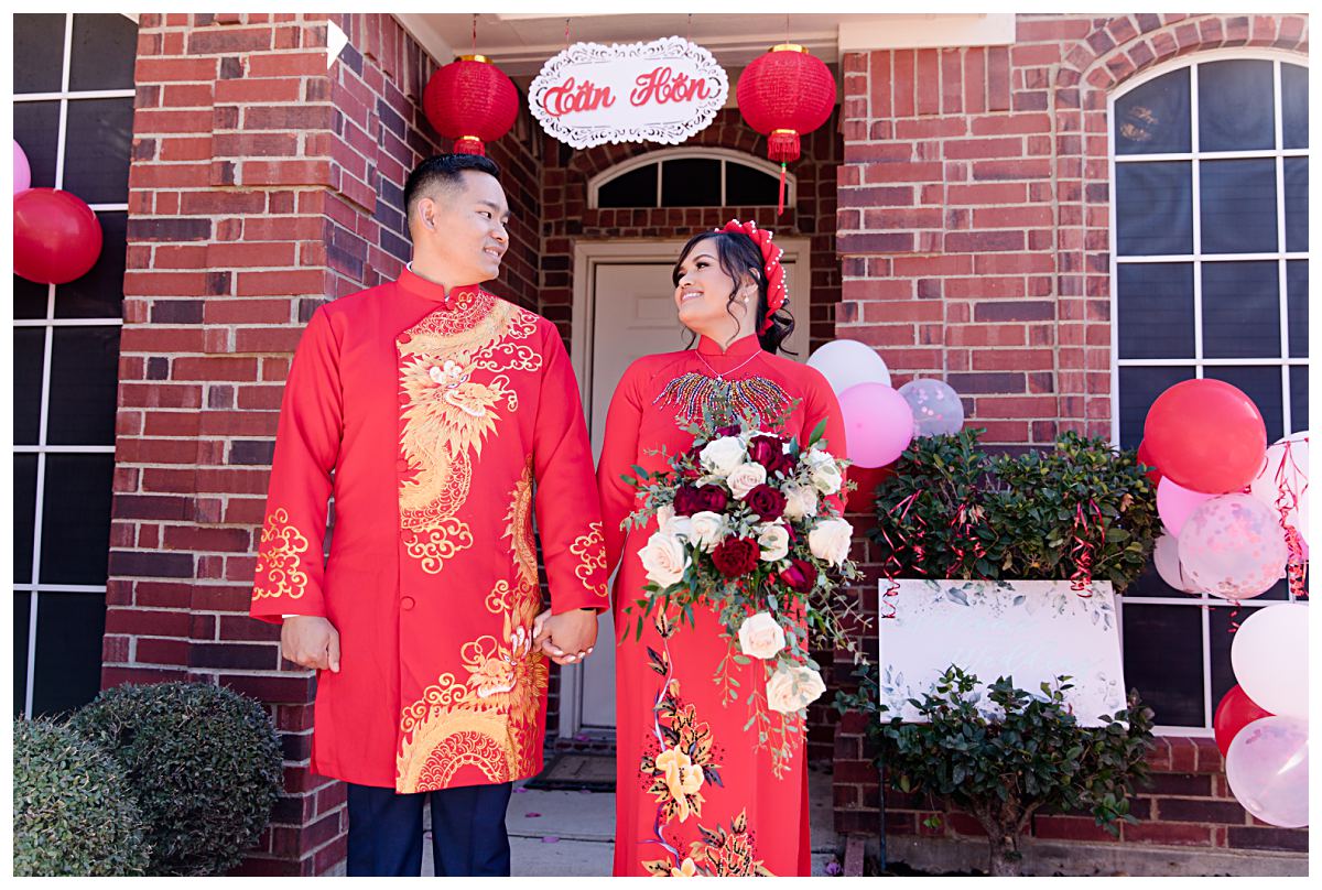 Bride and groom in traditional red ao dai smiling at one another in front of Tan Hon sign during Dallas Vietnamese tea ceremony photographed by Jenny Bui of Picture Bouquet Studio. 