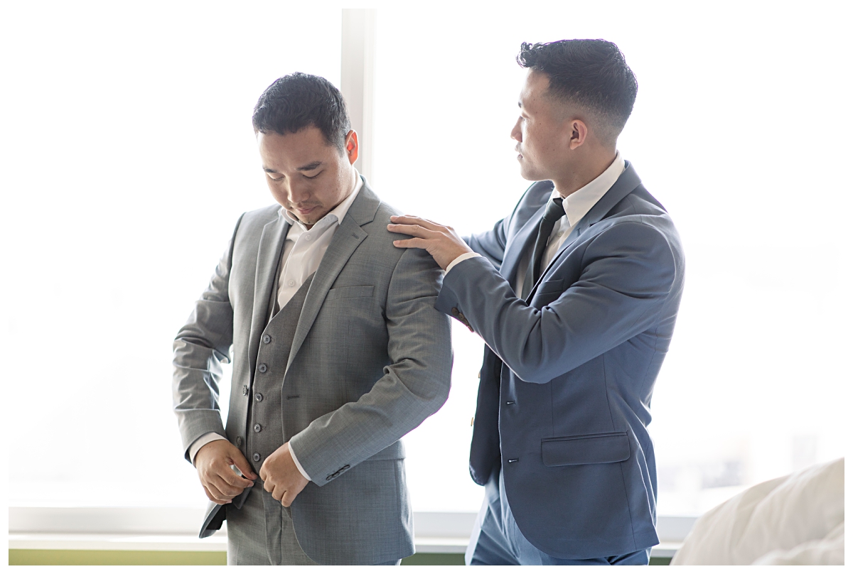 Groommen in blue suit helping groom in grey suit getting ready for wedding photographed by Dallas wedding photographer Jenny Bui of Picture Bouquet Studio. 