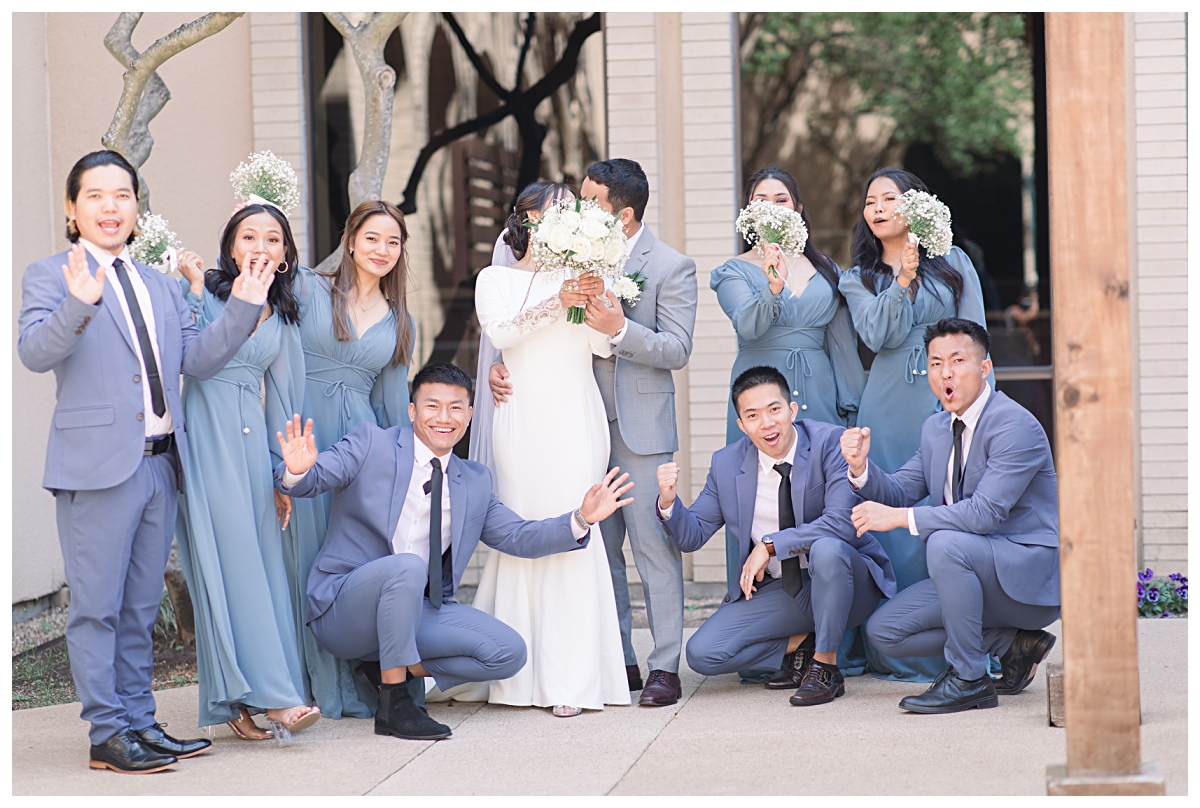 Bridal party in soft blue color palette cheers as they look at the camera and the bride and groom hides behind white rose bouquet for a kiss during wedding day at Highland Oaks Church of Christ from a Dallas Asian wedding photographed by Jenny Bui of Picture Bouquet Studio. 