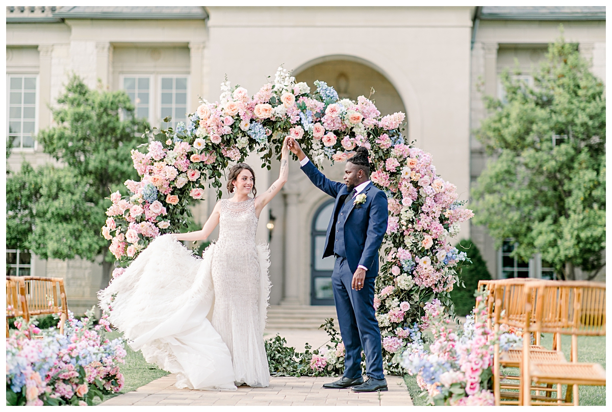 Groom in navy suit twirling bride in vintage feather wedding gown in front of floral circle at The Olana Wedding venue for styled shoot photographed by Dallas Wedding Photographer Jenny Bui of Picture Bouquet Studio. 