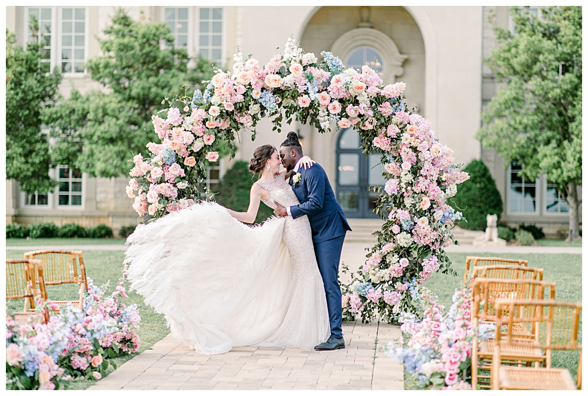 Groom in navy suit and bride in gorgeous feathered wedding gown leans in for kiss in front of pastel circle floral arch at The Olana Wedding venue for styled shoot photographed by Dallas Wedding Photographer Jenny Bui of Picture Bouquet Studio. 