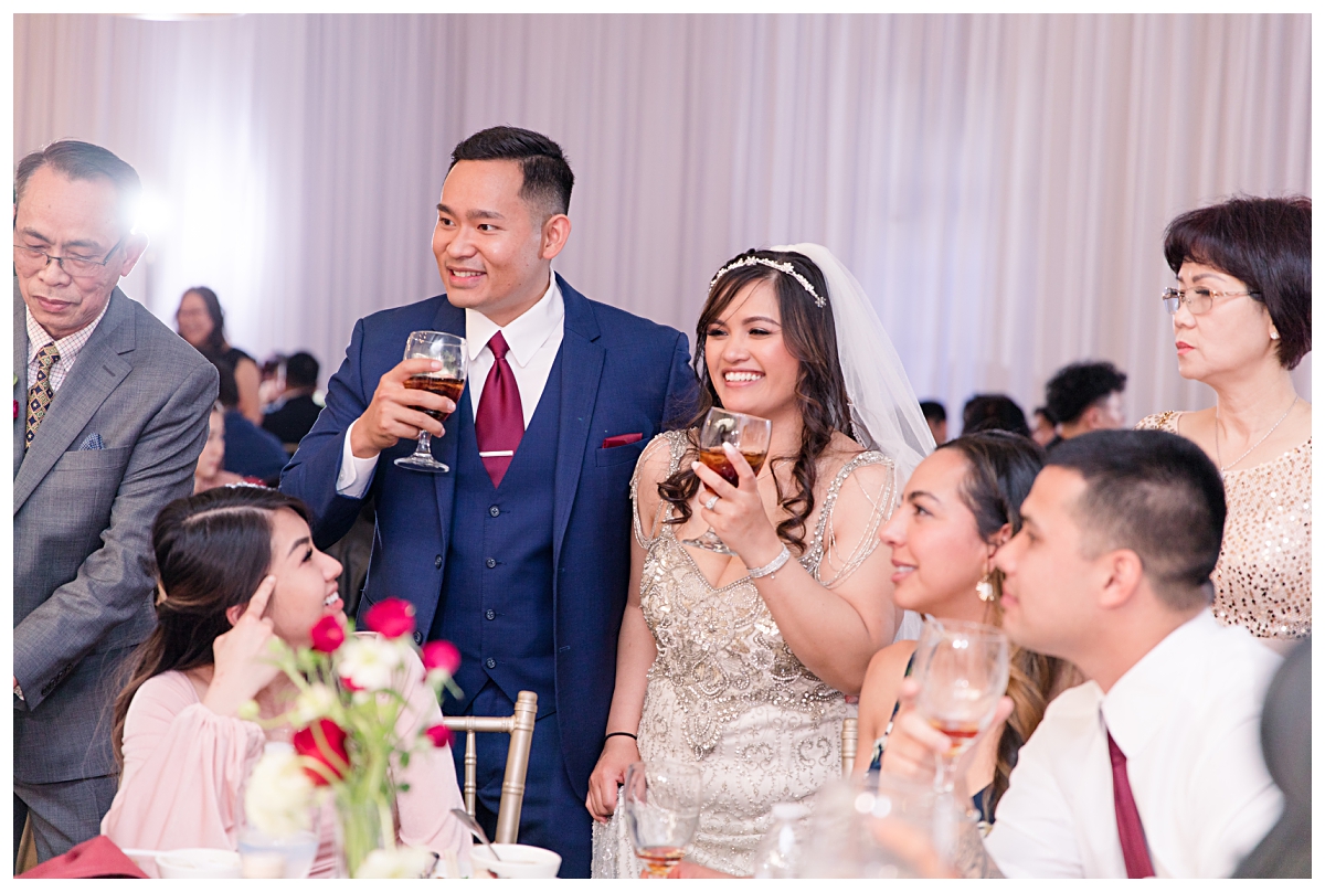Bride in vintage beaded wedding dress and groom in navy suit and burgundy tie greets and cheers with guests during Vietnamese table greetings at Zander House wedding photographed by Dallas wedding photographer Jenny Bui of Picture Bouquet Studio. 