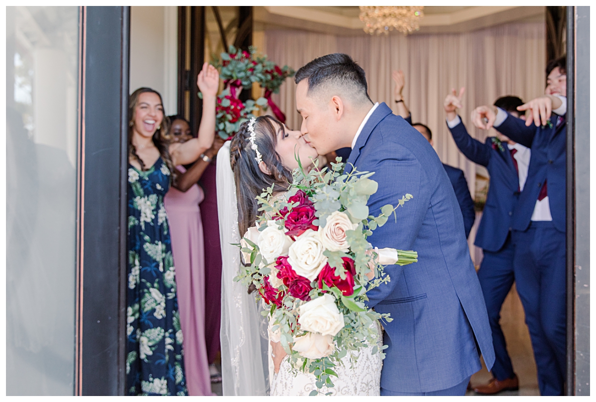Bride and groom shares kiss at end of exit as bridal party cheers in background during Dallas Vietnamese wedding at Zander House photographed by Dallas wedding photographer Jenny Bui of Picture Bouquet Studio. 