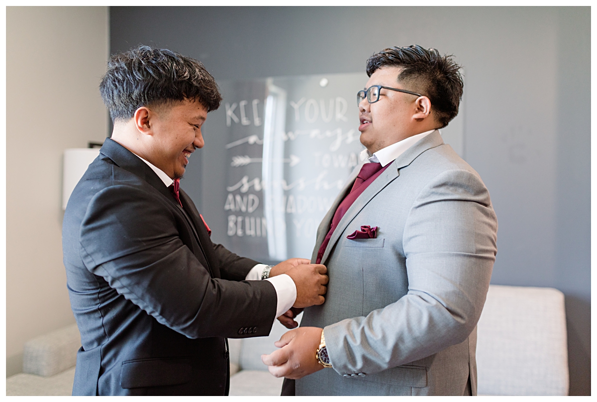 Groomsmen helping groom in burgundy vest and tie during getting ready portion of wedding day photographed by Dallas wedding photographer Jenny Bui of Picture Bouquet Studio. 