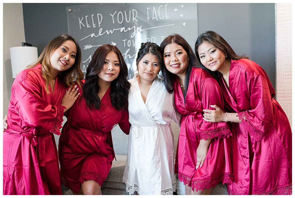 Bride in white robe poses with bridesmaids in matching burgundy robes for portraits during getting ready portion of wedding day photographed by Dallas wedding photographer Jenny Bui of Picture Bouquet Studio. 