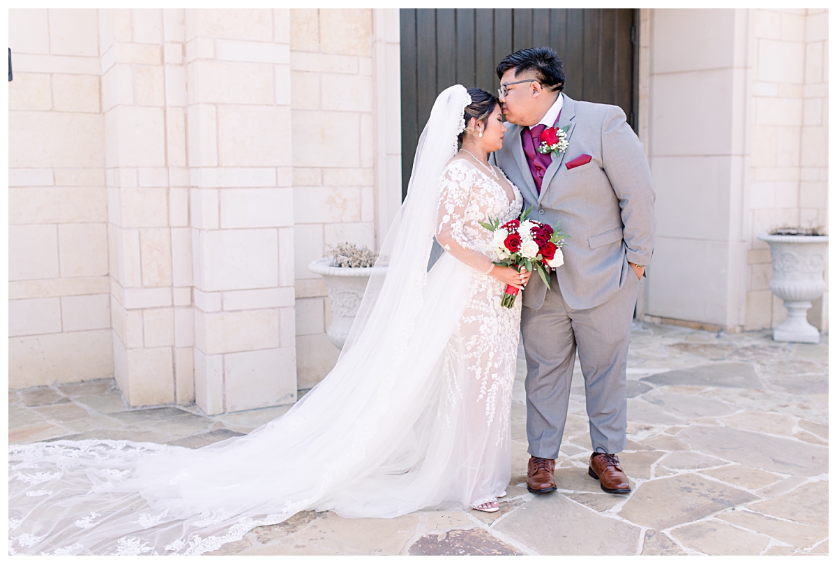 Groom kissing bride on forehead in front of Riverwalk Chapel for wedding portraits at Flower Mound Riverwalk photographed by Dallas wedding photographer Jenny Bui of Picture Bouquet Studio. 