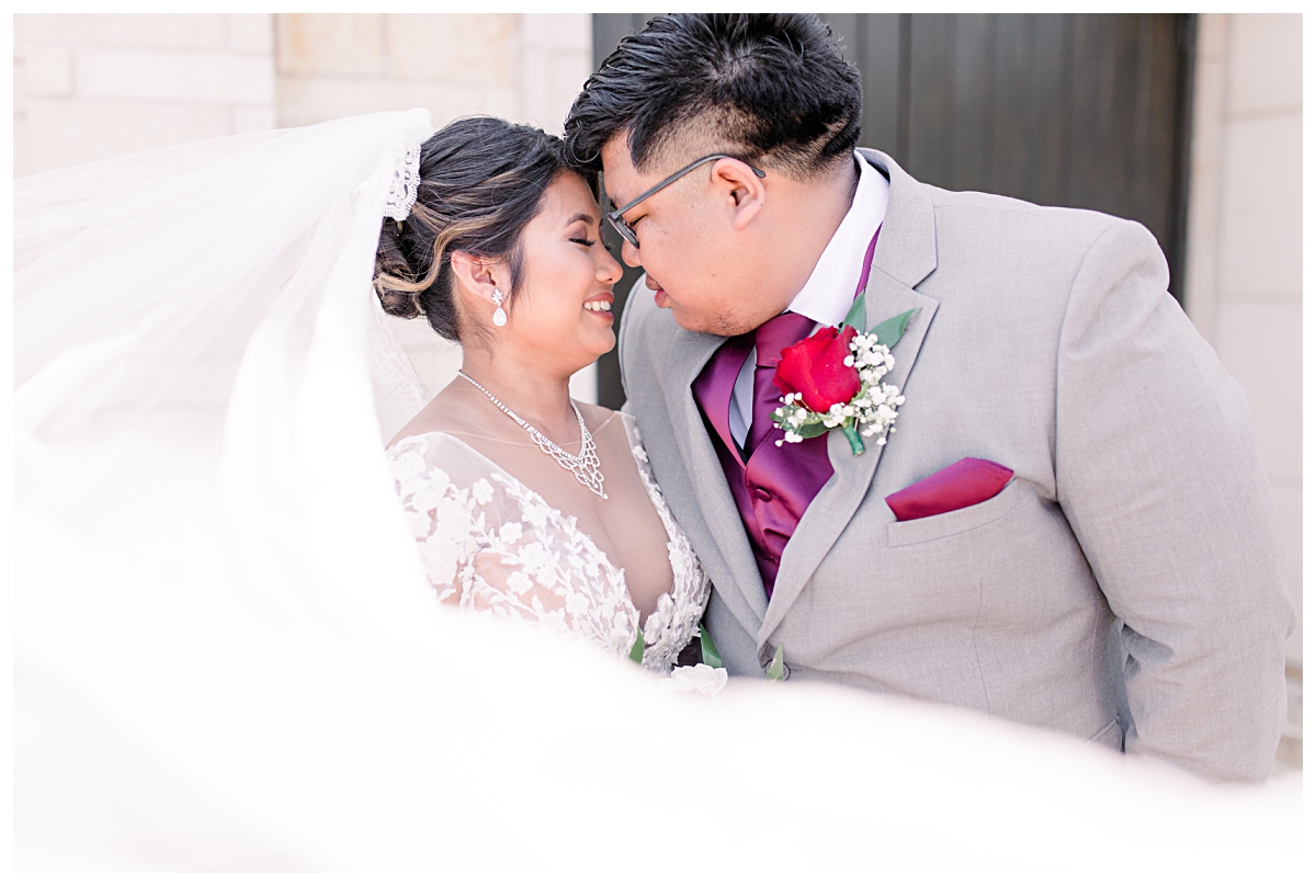 Gorgeous Asian bride in white lace wedding dress and groom in light gray suit with burgundy vest and tie leans into one another with veil wrapped around them for wedding portraits at Flowermound Riverwalk photographed by Dallas wedding photographer Jenny Bui of Picture Bouquet Studio. 