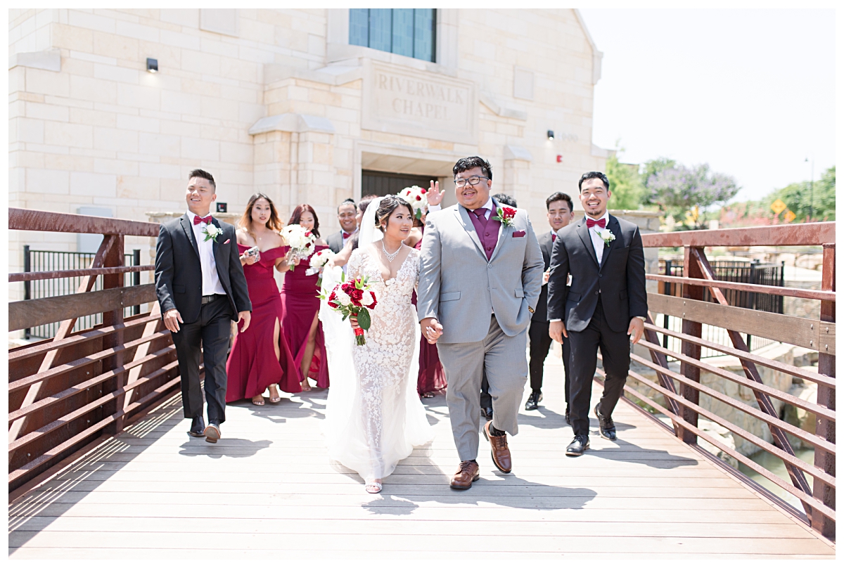 Bride and groom walks across bridge at Riverwalk Chapel in Lewisville Texas with bridal party in burgundy and cream for Lewisville Texas wedding for wedding portraits at Flowermound Riverwalk photographed by Dallas wedding photographer Jenny Bui of Picture Bouquet Studio. 