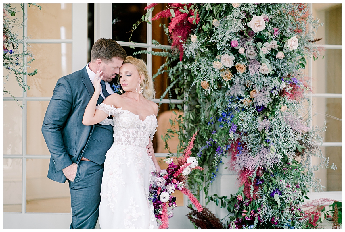 Bride in off shoulder white wedding dress caressing groom's face with her hands by floral arches in front of glass doorway at The Olana photographed by Dallas wedding photographer Jenny Bui of Picture Bouquet Studio. 