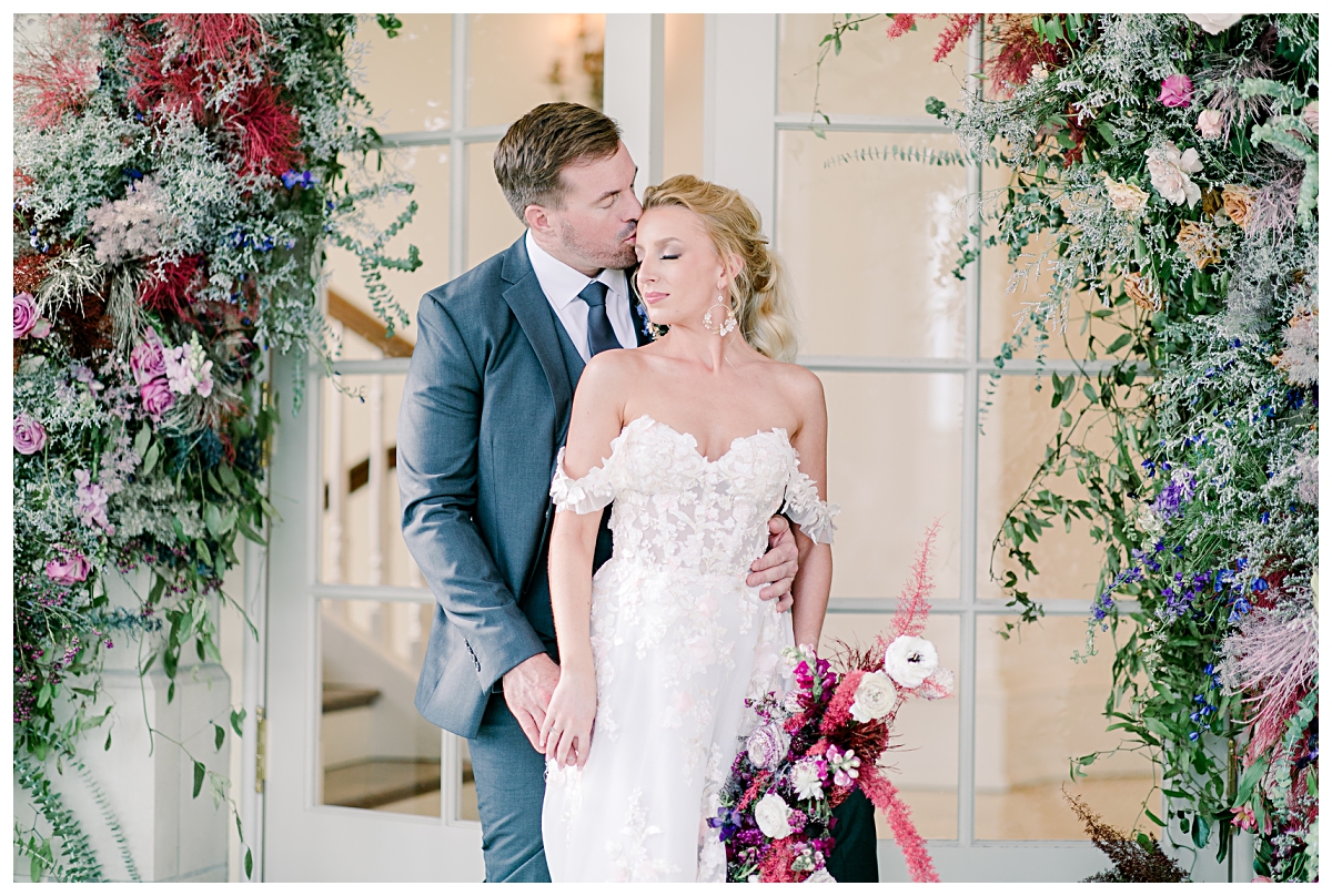Groom kissing bride in off shoulder wedding dress by flower arches in front of glass doorway at The Olana photographed by Dallas wedding photographer Jenny Bui of Picture Bouquet Studio. 