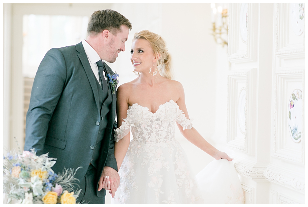 Bride in off shoulder white lace wedding dress laughing and talking with groom in blue suit as they walk down the hallways of one of the best dallas wedding venue, The Olana, photographed by Jenny Bui of Picture Bouquet Studio. 