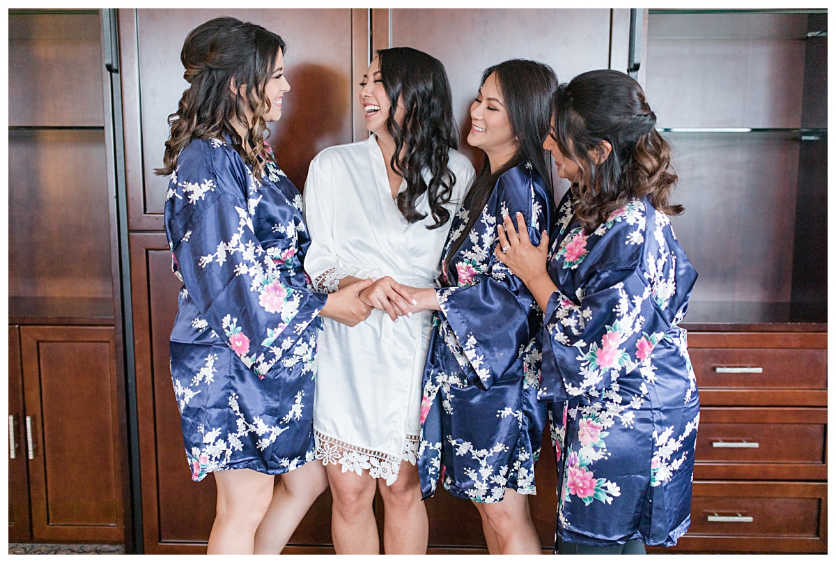 Bride laughing with bridesmaids in navy and floral robe during getting ready portion on wedding day photographed by Dallas wedding photographer Jenny Bui of Picture Bouquet Studio for Holy Trinity Catholic Church wedding in Dallas, TX.  