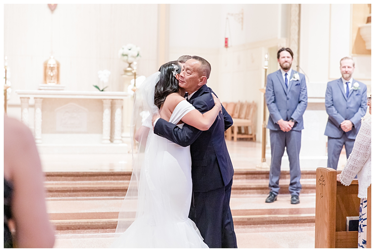 Father of the bride gives her a hug before taking a seat during wedding ceremony photographed by Dallas wedding photographer Jenny Bui of Picture Bouquet Studio for Holy Trinity Catholic Church wedding in Dallas, TX.  