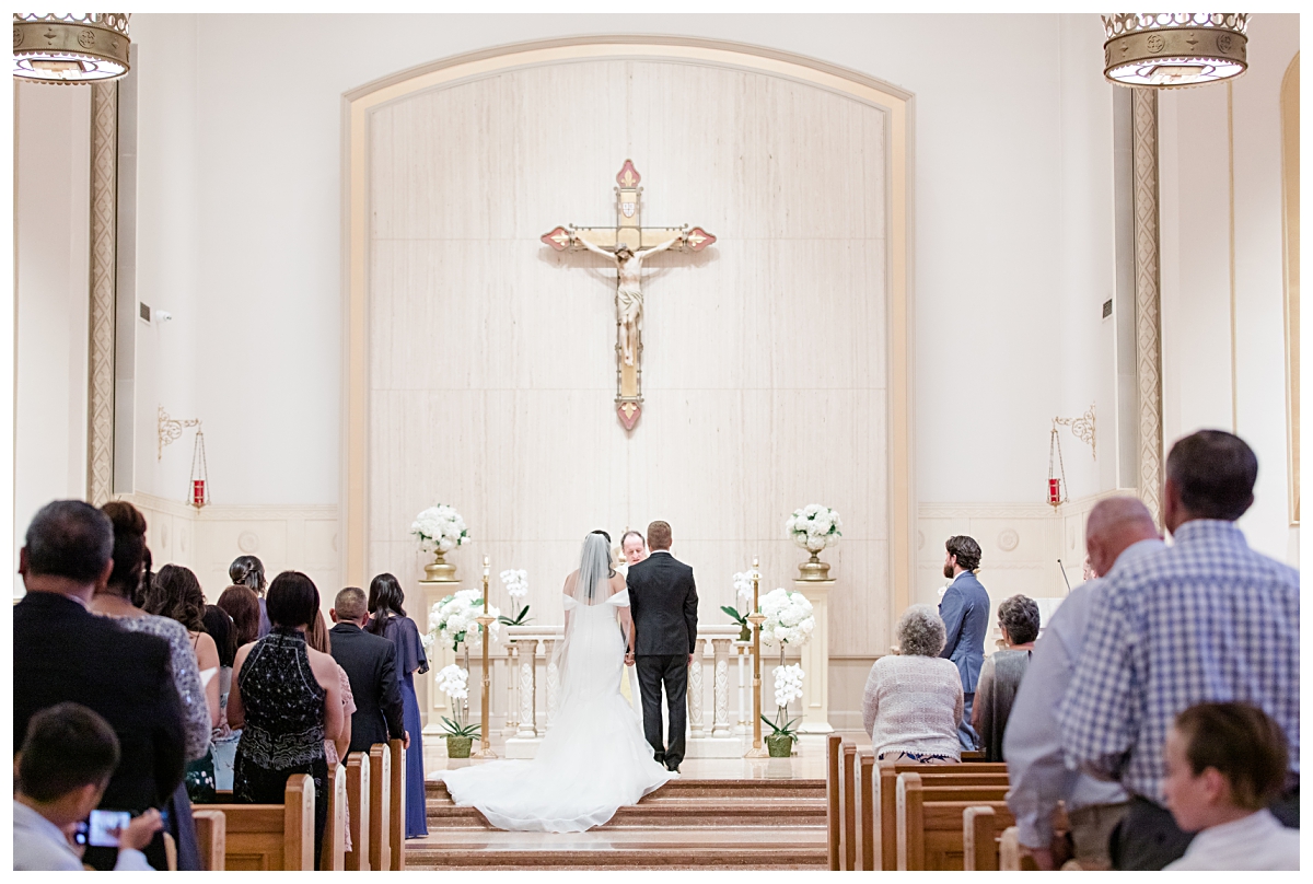 Back view of bride and groom in front of priest and guests during wedding day ceremony photographed by Dallas wedding photographer Jenny Bui of Picture Bouquet Studio for Holy Trinity Catholic Church wedding in Dallas, TX.  