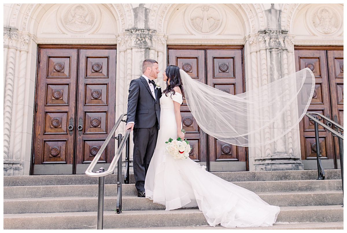 Bride and groom in front of Holy Trinity Catholic Church with floating veil behind her photographed by Dallas wedding photographer Jenny Bui of Picture Bouquet Studio for Holy Trinity Catholic Church wedding in Dallas, TX.  