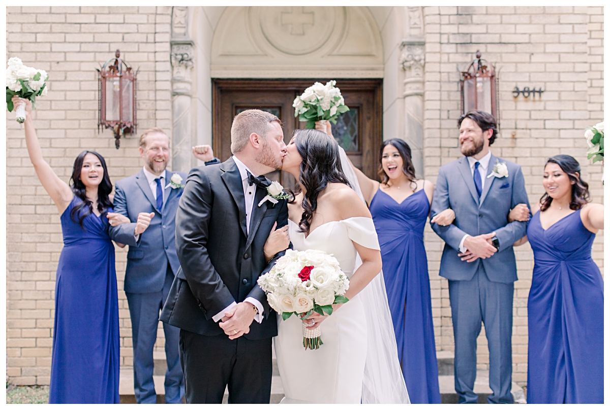 Bride and groom kisses as bridal party cheers in background photographed by Dallas wedding photographer Jenny Bui of Picture Bouquet Studio for Holy Trinity Catholic Church wedding in Dallas, TX.  