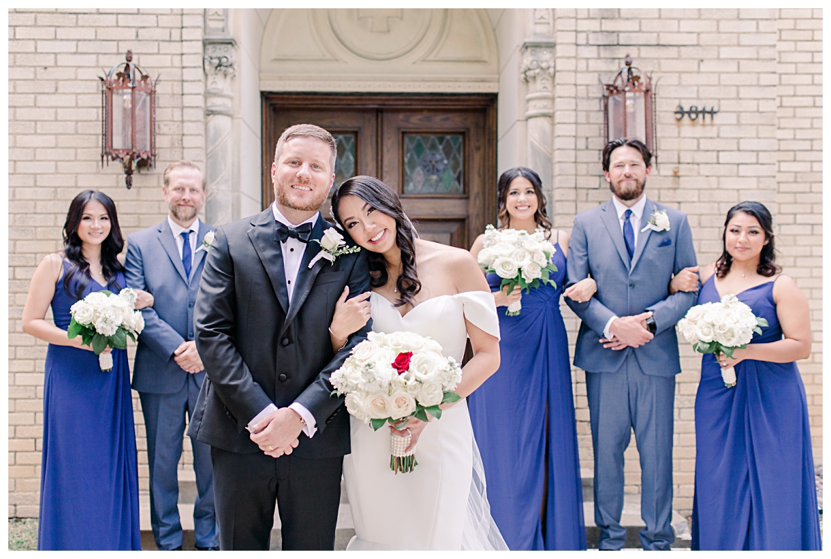 Beautiful bride leaning on groom as royal blue bridal party stands in background photographed by Dallas wedding photographer Jenny Bui of Picture Bouquet Studio for Holy Trinity Catholic Church wedding in Dallas, TX.  