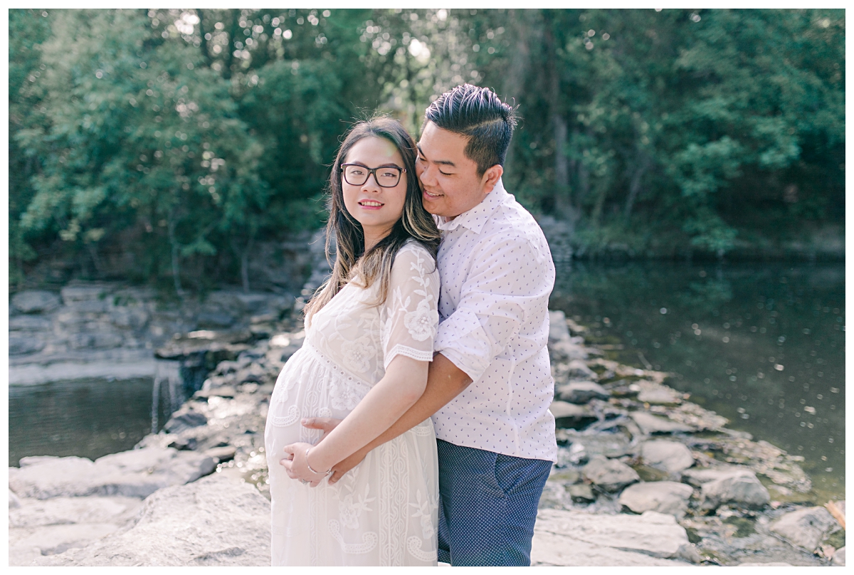 Husband hugging wife in long white maternity gown for for Prairie Creek Maternity session photographed by Dallas wedding and portrait photographer Jenny Bui of Picture Bouquet Studio. 