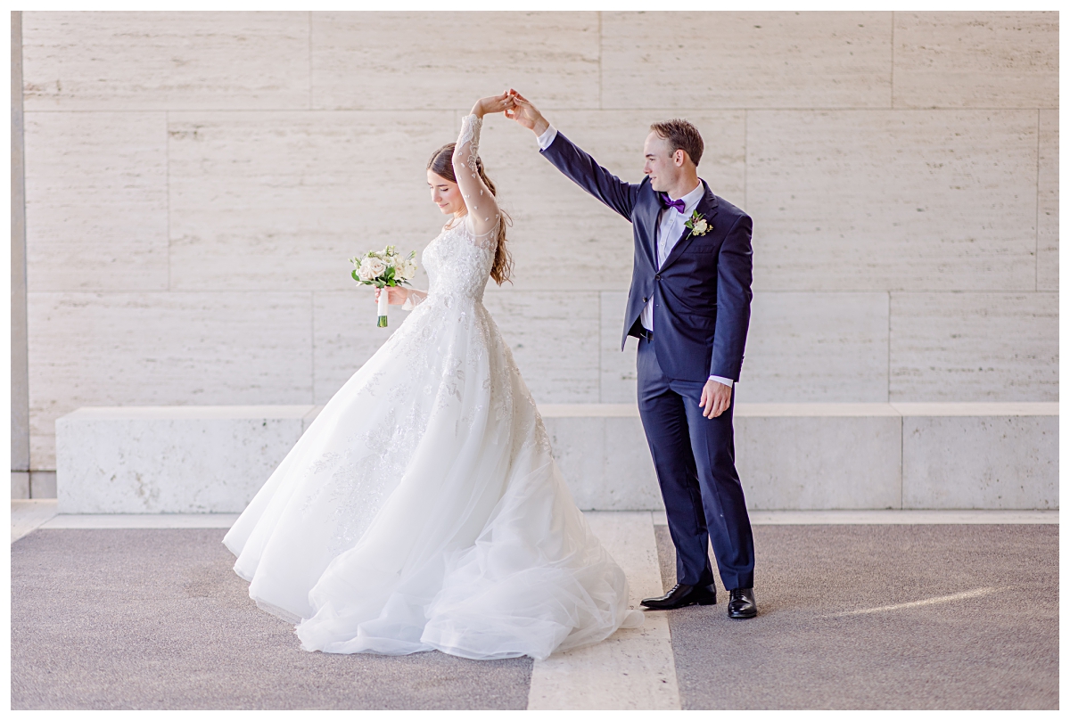 Groom in navy suit twirls his beautiful bride in a long sleeve white lace wedding gown for Kimbell Art Museum wedding portraits photographed by Dallas wedding photographer Jenny Bui of Picture Bouquet Studio. 