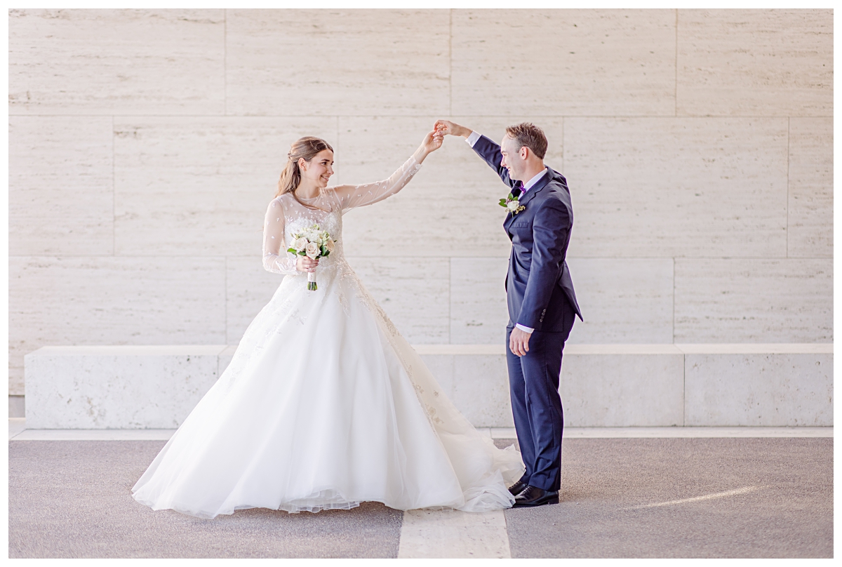 Groom in navy suit twirling his beautiful bride in a long sleeve white lace wedding gown for Kimbell Art Museum wedding portraits photographed by Dallas wedding photographer Jenny Bui of Picture Bouquet Studio. 