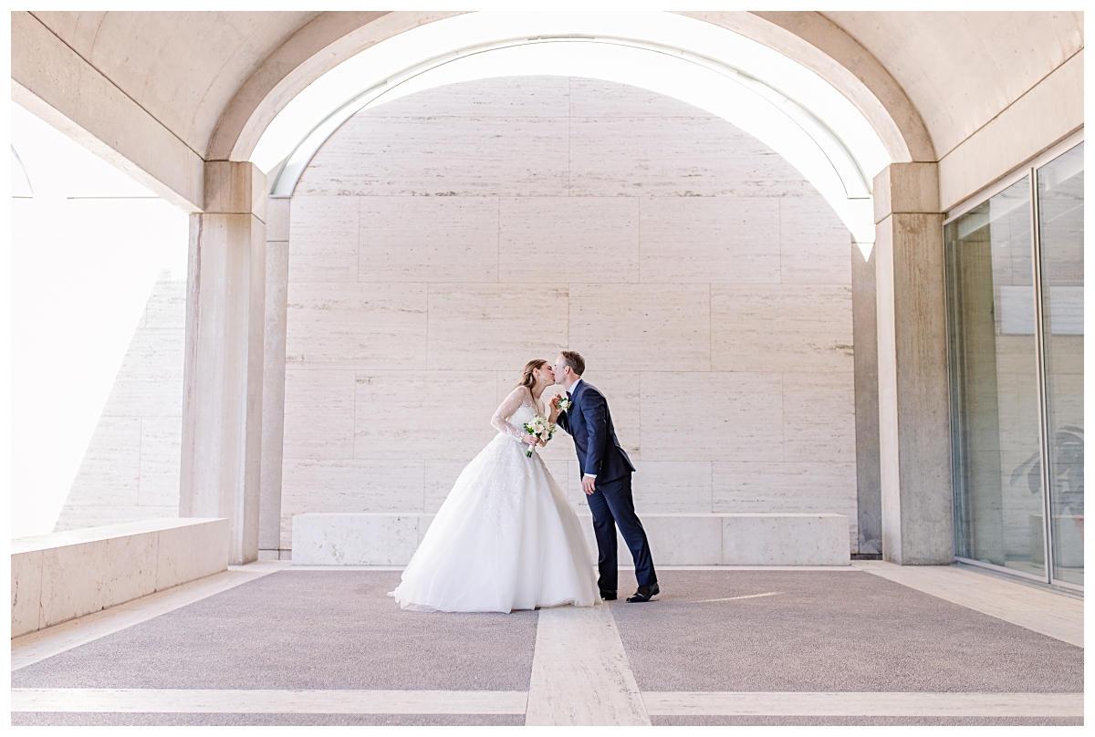 Bride in white wedding dress leans in for a kiss with her groom in navy suit underneath archway for Kimbell Art Museum wedding portraits photographed by Dallas wedding photographer Jenny Bui of Picture Bouquet Studio. 