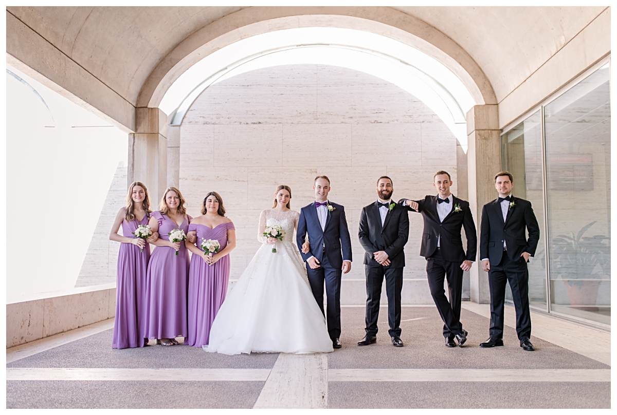 Bridal party in lilac colors poses with bride in long sleeve lace wedding dress and groom in navy suit under archway for Kimbell Art Museum wedding portraits photographed by Dallas wedding photographer Jenny Bui of Picture Bouquet Studio. 
