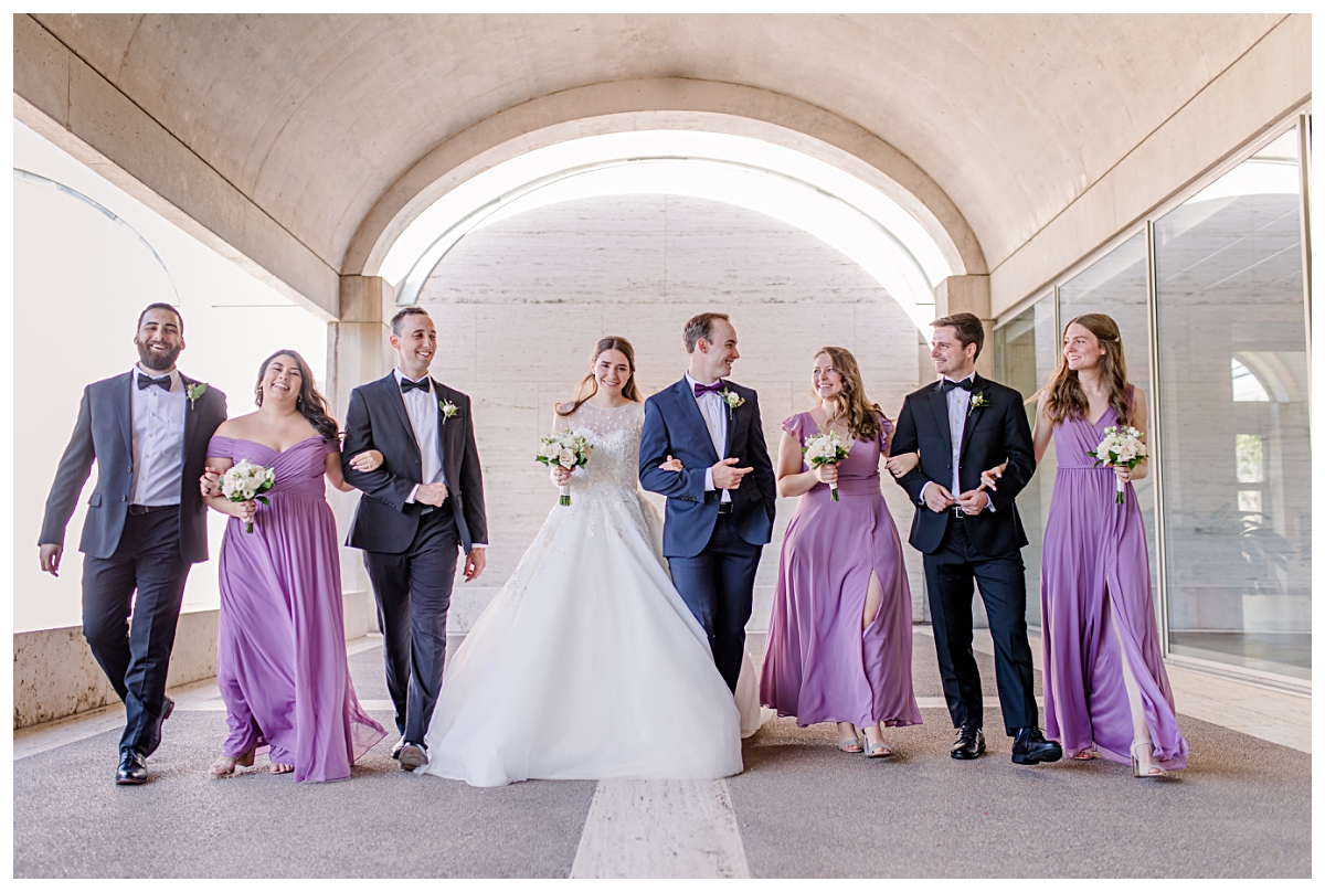 Wedding party in lilac colors walking and laughing underneath archway at Kimbell Art Museum for wedding day portraits photographed by Jenny Bui of Picture Bouquet Studio. 