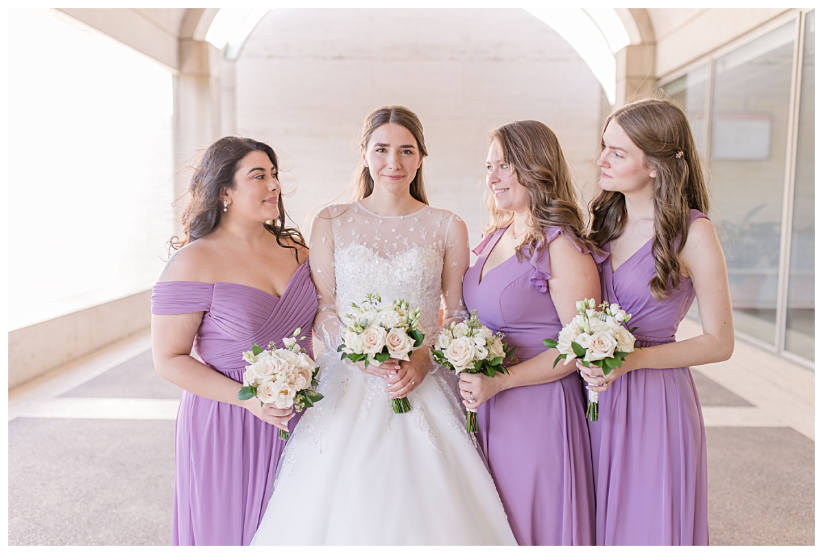 Bridesmaids in lilac dresses smiles at gorgeous bride in long sleeve white lace wedding dress at Kimbell Art Museum for wedding day portraits photographed by Jenny Bui of Picture Bouquet Studio. 